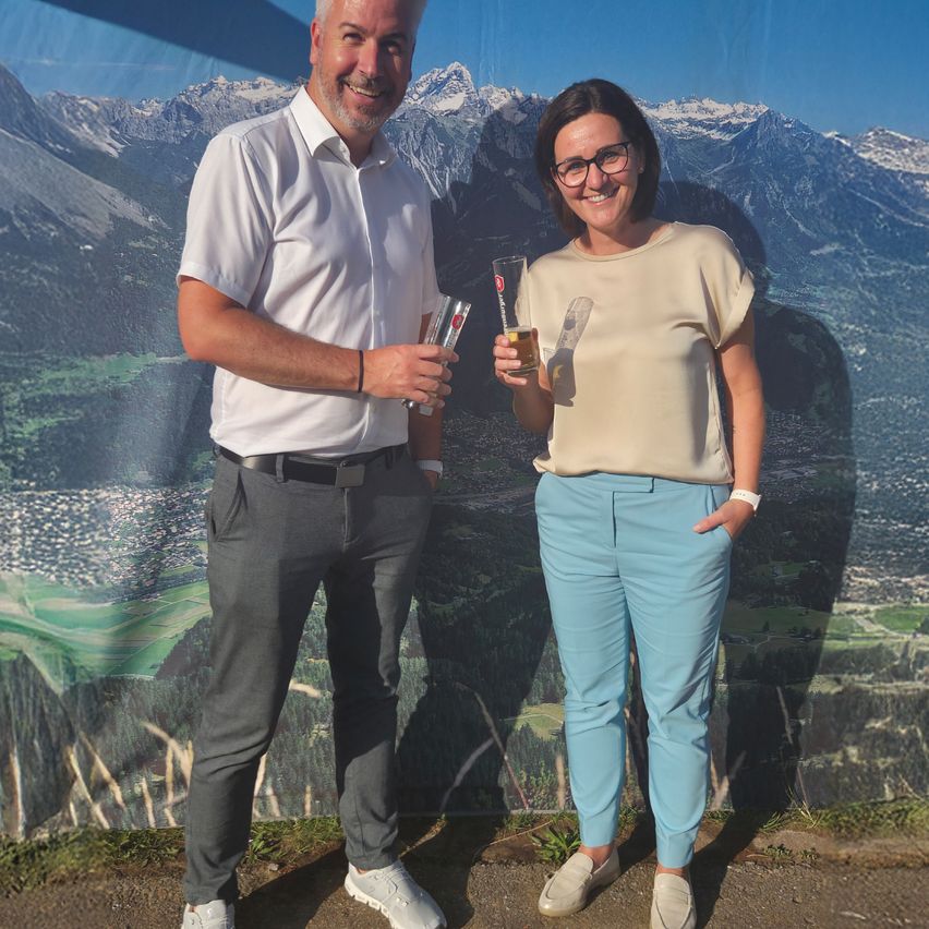 Ein Mann und eine Frau stehen vor einer Bergkulisse, halten Biergläser in der Hand und lächeln beide.