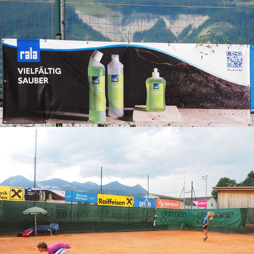 Auf einem Tennisplatz im Freien ist eine große Werbung für 'Rala'-Reinigungsprodukte zu sehen, bei der zwei Flaschen abgebildet sind. Unten sind Spieler in einem Tennismatch zu sehen, mit Bergen im Hintergrund.