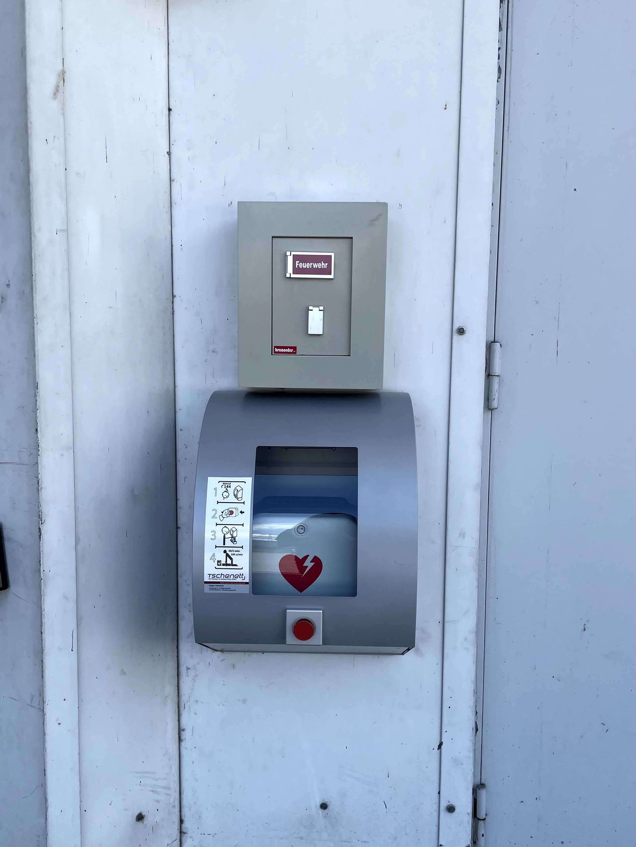 Ein Feuerlöscher ist an einer weißen Wand montiert. Er hat eine graue Metallbox mit einem Schalter darüber. Darunter befindet sich eine graue Maschine mit einem roten Herzsymbol und einem Aufkleber mit Zahlen und Symbolen.