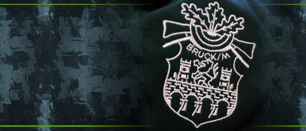 Eine Nahaufnahme eines schwarzen Kleidungsstücks zeigt ein gesticktes Logo mit der Aufschrift 'BRUCK/IN'. Das Logo zeigt eine Brücke und eine Burg mit einem Pferd und Reiter, umgeben von Blättern.