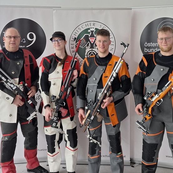 Vier Personen, gekleidet in passenden Uniformen, posieren mit ihren Gewehren vor einer Kulisse mit Logos und der Nummer 9.