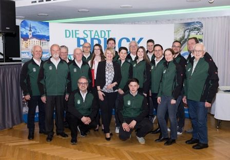 Eine Gruppe von Menschen in grünen Jacken posiert für ein Foto in einem Raum. Das Banner steht für 'Die Stadt Bruck'.