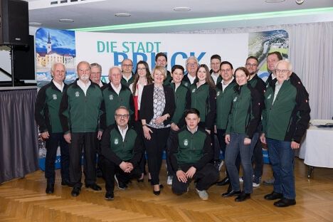Eine Gruppe von Menschen in grünen Jacken posiert für ein Foto in einem Raum. Das Banner steht für 'Die Stadt Bruck'.