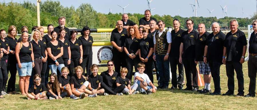 Eine Gruppe von Menschen in passenden schwarzen T-Shirts mit einem Logo auf der Rückseite posiert für ein Foto in einer Outdoor-Umgebung. Einige tragen Brillen, Armbänder und Turnschuhe. Hinter ihnen ist eine gelbe Barriere mit einem weißen Schild. In der Ferne gibt es Bäume und Windräder.