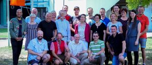 Eine Gruppe von Menschen posiert für ein Foto vor einem Gebäude mit der Aufschrift 'Musikschule'. Die Gruppe besteht aus Männern und Frauen, die alle lässig gekleidet sind. Einige lächeln und scheinen den Moment zu genießen.