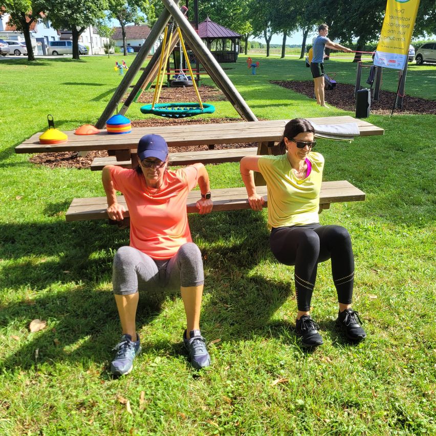 Zwei Frauen machen Sit-ups auf einer Bank im Park. Hinter ihnen befindet sich ein Tisch, Spielzeug und eine Schaukel. Ein Mann spielt im Hintergrund mit einem Hund.