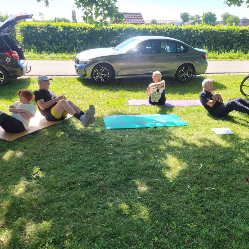 Vier Personen machen Yoga auf dem Rasen. Ein Auto ist hinter ihnen geparkt, und ein Fahrrad steht rechts. Sie haben Matten und Papier auf dem Boden.