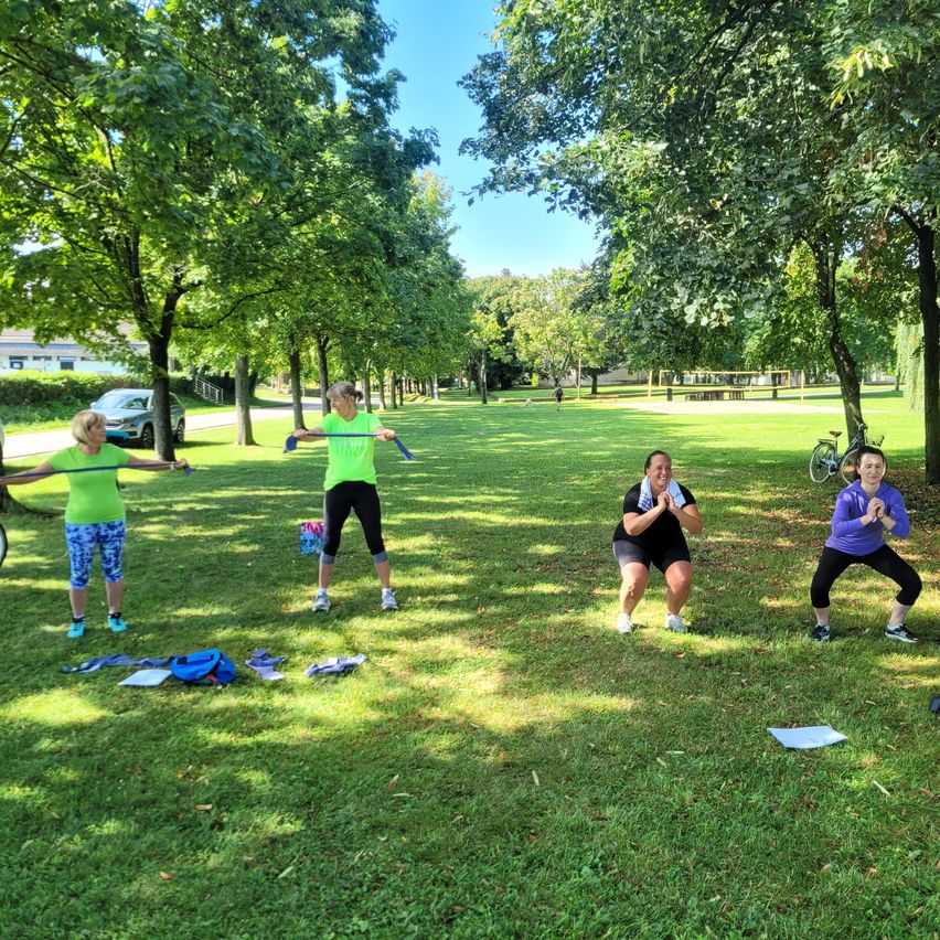 In einem Park führen eine Gruppe von Frauen Übungen mit Widerstandsbändern durch. Einige stehen, andere knien. Ein Fahrrad ist in der Nähe der Bäume geparkt.
