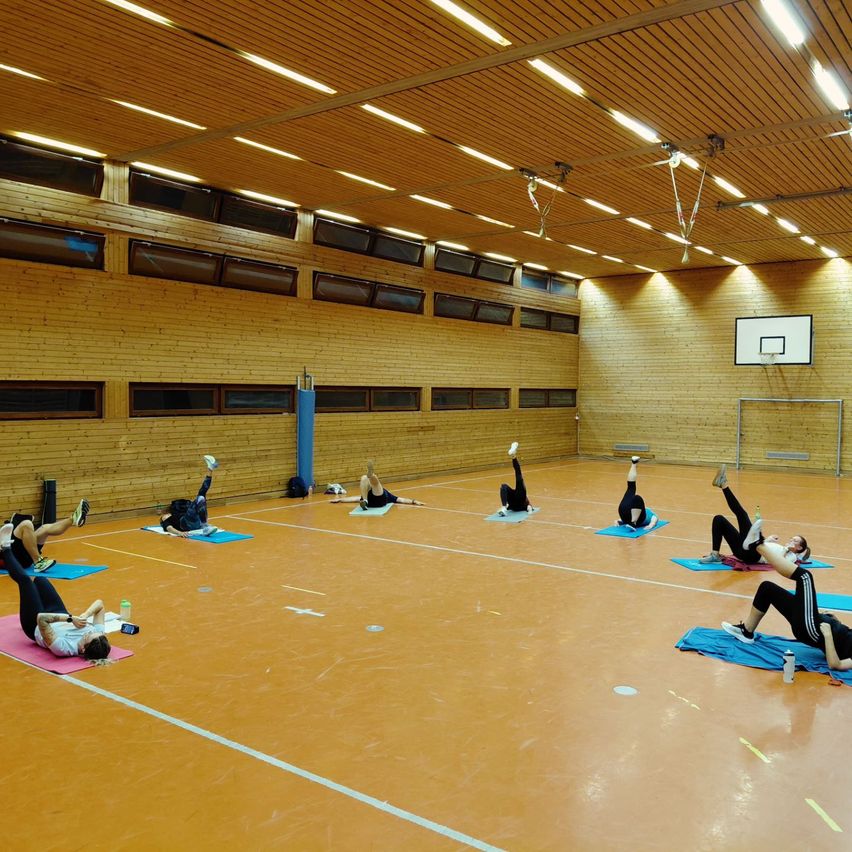 In einer Sporthalle üben mehrere Personen Yoga auf blauen Matten. Holzwände und Decke mit Beleuchtung im Hintergrund. Ein Basketballkorb ist auf der rechten Seite sichtbar.