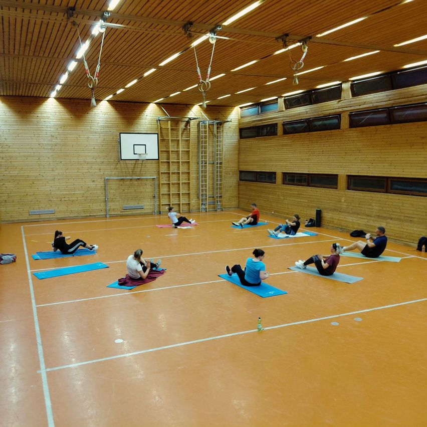 Ein Indoor-Fitnessstudio mit Holzwänden verfügt über einen Basketballkorb. Mehrere Personen praktizieren Yoga auf blauen Matten. Der Boden ist mit weißen Linien markiert.