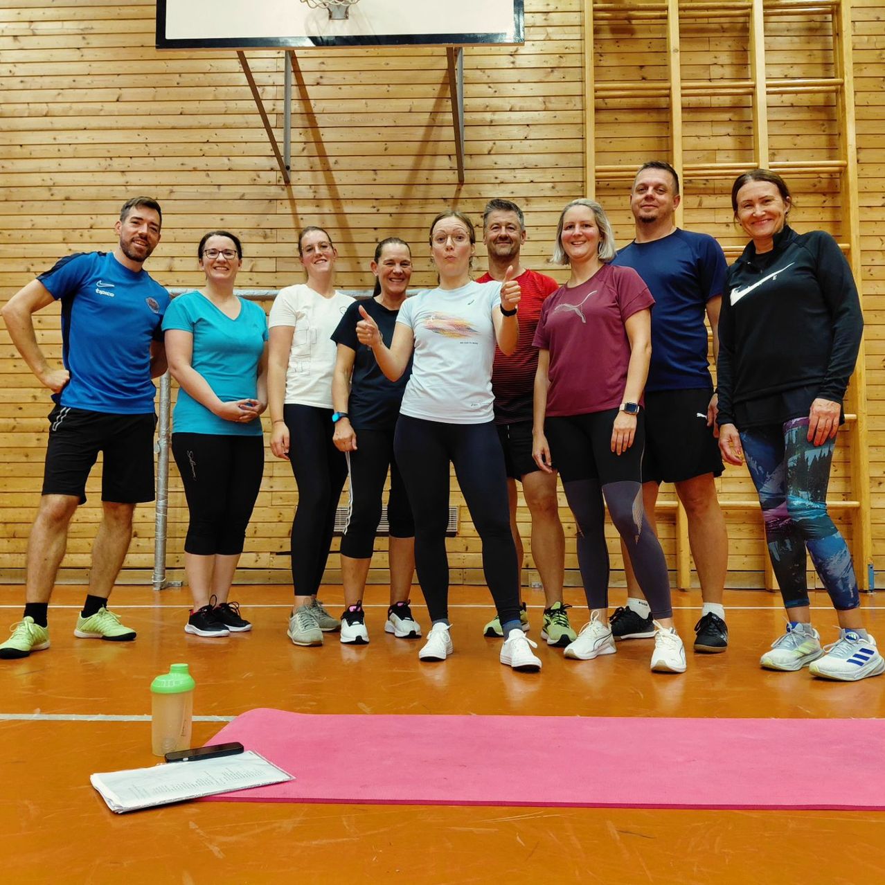 Gruppe von Menschen in einem Fitnessstudio, die in zwei Reihen stehen, wobei eine Frau vorne den Daumen hochhält. Holzwände und ein Basketballkorb im Hintergrund.