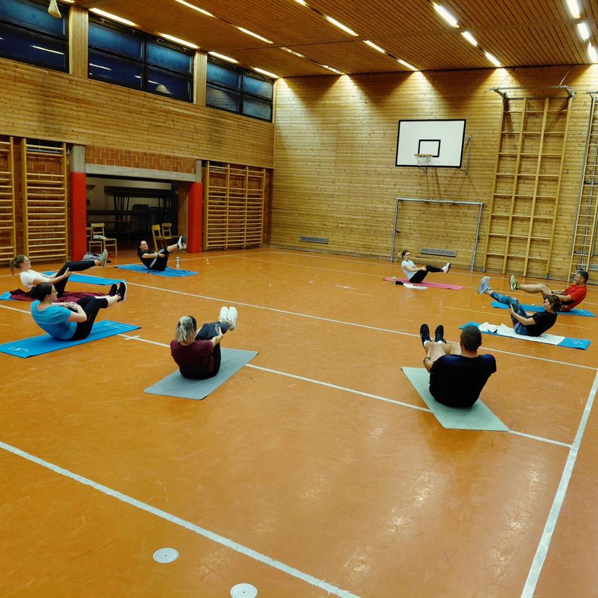 In einer Turnhalle macht eine Gruppe von Menschen Yoga auf blauen Matten. Die Turnhalle hat Holzwände, Fenster und einen Basketballkorb. Sie tragen Freizeitkleidung und Turnschuhe.