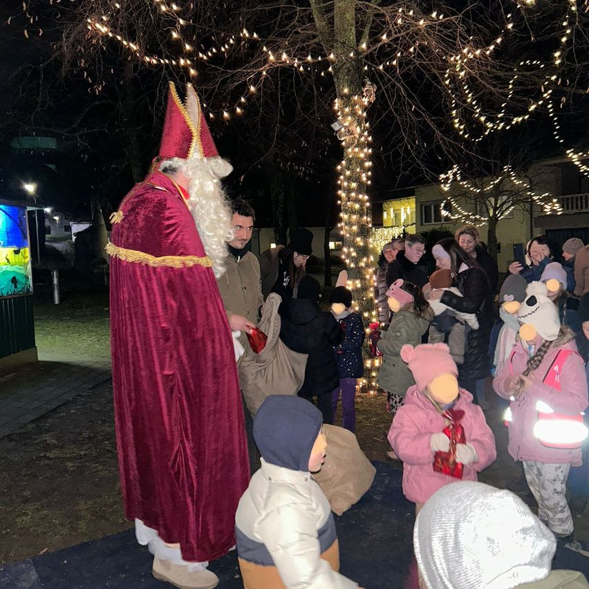 Ein Mann in Santa Claus-Kostüm verteilt Geschenke an Kinder unter einem dekorierten Baum bei Nacht.