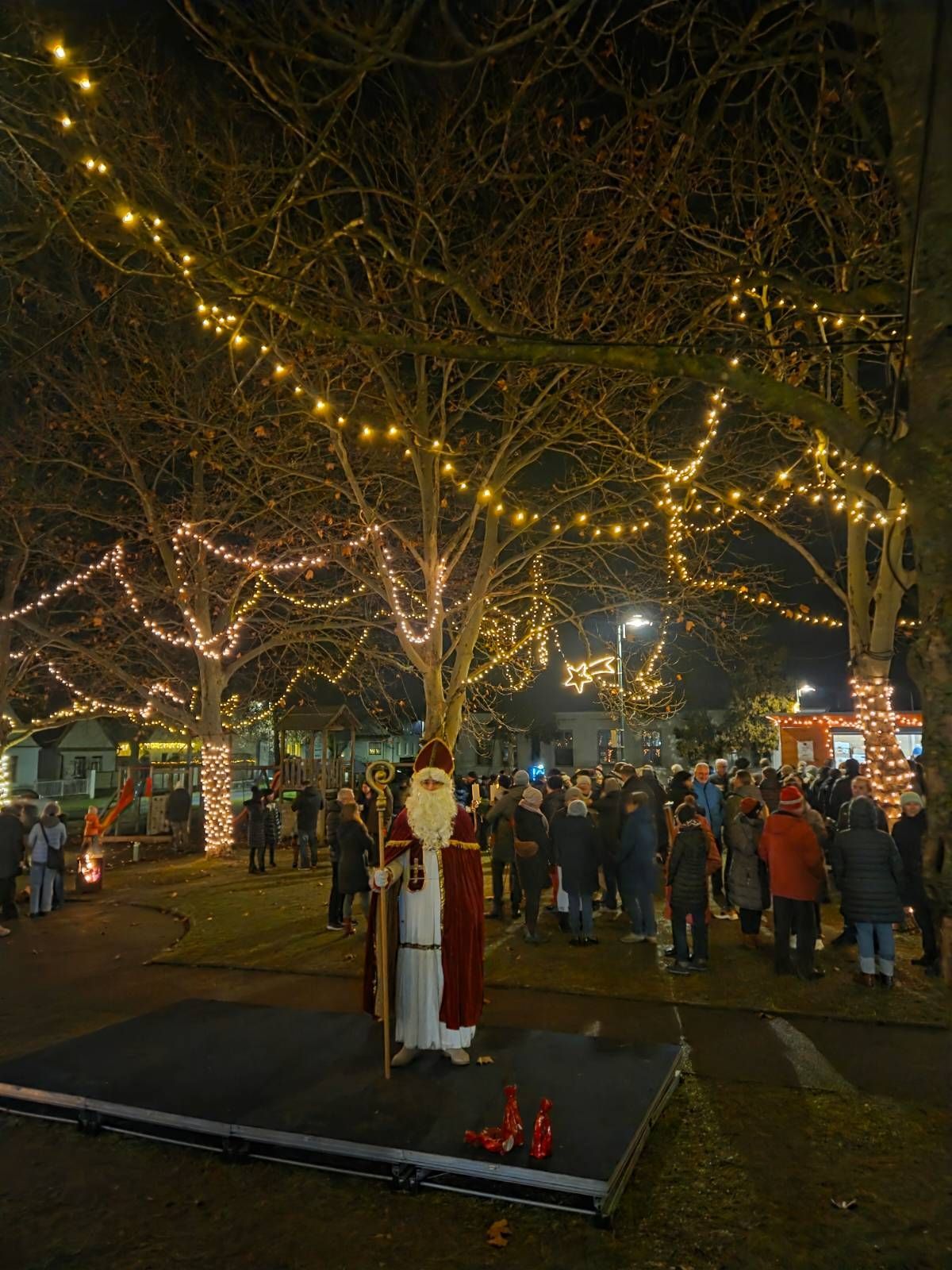 Eine Nachtszene zeigt einen Mann, der als Weihnachtsmann verkleidet ist und auf einer Bühne steht, umgeben von einer Menschenmenge. Das Gebiet ist mit Weihnachtslichtern an Bäumen und Gebäuden geschmückt, und in der Nähe befindet sich ein Feuerkorb.