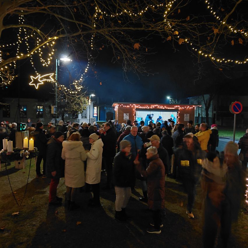 Eine Menschenmenge versammelt sich am Abend im Freien. Es gibt beleuchtete Bäume, eine Anzeige mit einem Bildschirm und Kerzen auf einem Tisch. Es scheint eine festliche Veranstaltung zu sein.