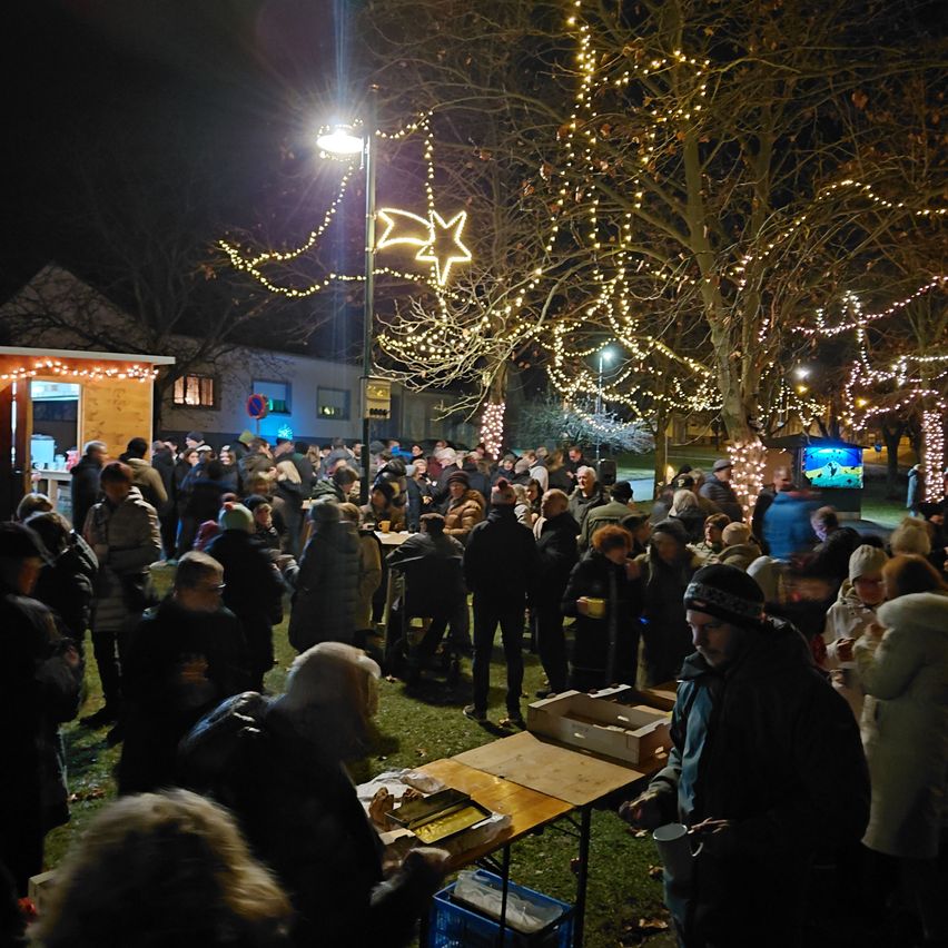 Eine Menschenmenge versammelt sich bei Nacht zu einer Veranstaltung im Freien. Sie stehen um Tische mit Essen, und einige trinken. Der Bereich ist mit Weihnachtsbeleuchtung und Bäumen geschmückt.