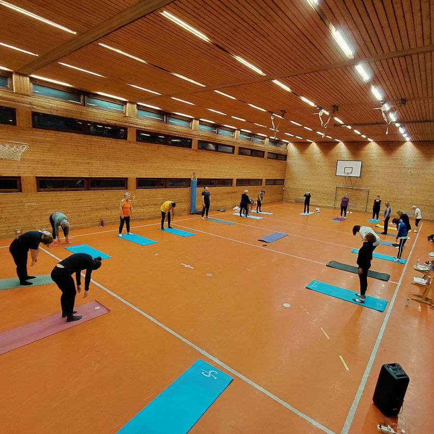 Innengymnasium mit mehreren Personen, die Yoga auf Matten üben. Holzwände und Decke, Basketballkorb in der Ecke, verstreute Matten.