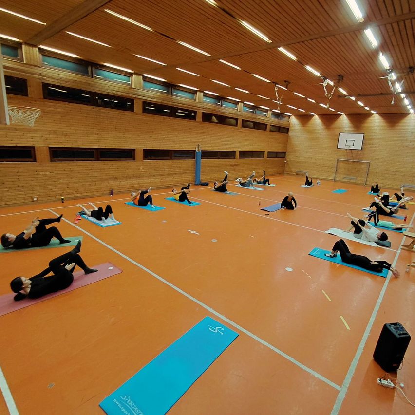 Eine Gruppe von Menschen macht Yoga auf Matten in einer Turnhalle. Die Turnhalle hat Holzwände und einen Basketballkorb.