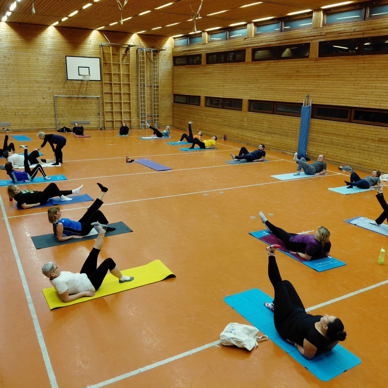 Ein Fitnessstudio mit Holzwänden und orangefarbenem Boden, wo Menschen auf Matten Yoga machen. Ein Basketballkorb ist an einer Wand montiert.