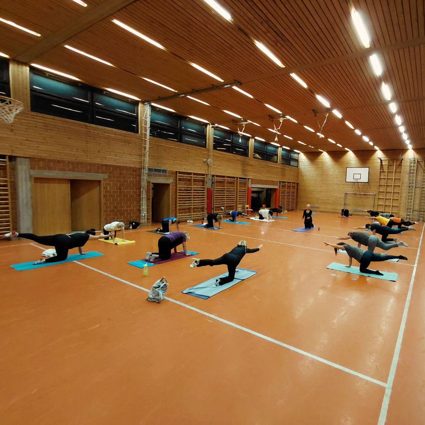 Eine Gruppe von Menschen in einer Turnhalle macht Yoga auf blauen Matten auf dem Boden. Einige befinden sich in verschiedenen Posen, während andere ruhen. Die Turnhalle hat Holzwände, einen Basketballkorb und Leuchtkörper an der Decke.
