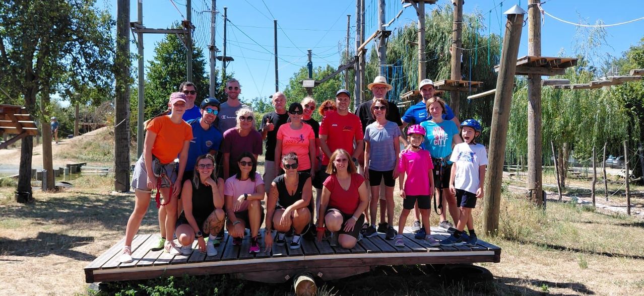 A group of people, including adults and children, are posing for a photo in front of an outdoor adventure course with climbing structures and trees in the background.