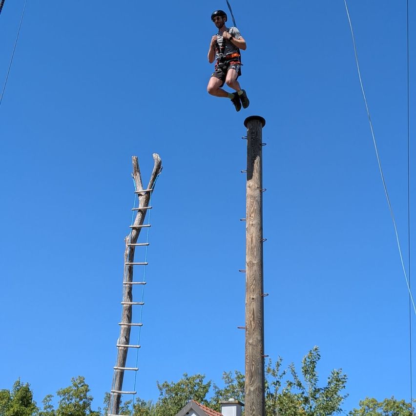 A person wearing a helmet and harness jumps from a rope attached to a tall wooden pole, with a ladder leaning against another pole nearby.