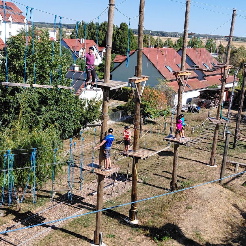 A group of people are navigating an outdoor obstacle course with wooden platforms and rope bridges. The course is surrounded by trees and houses.