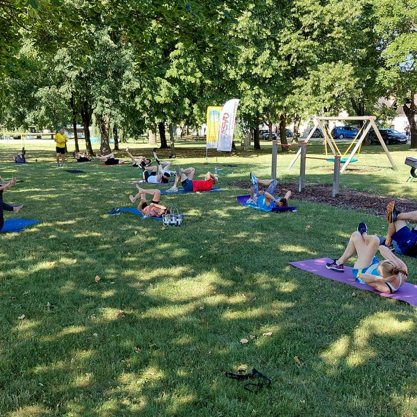 A group of people are doing yoga on the grass in a park with trees and a banner.