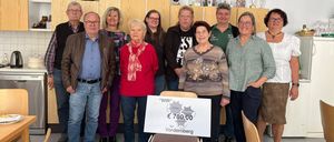 Eine Gruppe von Menschen posiert für ein Foto mit einem Schild, das 'WIR' und 'E 700,00 für Vordernberg' zeigt. Die Menschen lächeln und stehen hinter einem Stuhl mit einem Schild.