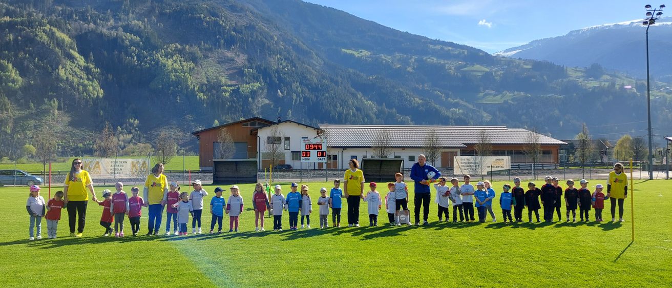 Eine Gruppe von Kindern und Erwachsenen steht auf einem Feld mit einer Bergkette im Hintergrund. Sie tragen verschiedenfarbige Shirts und Mützen.