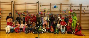A group of children in costumes pose for a picture in a gym. There are balloons and decorations behind them.