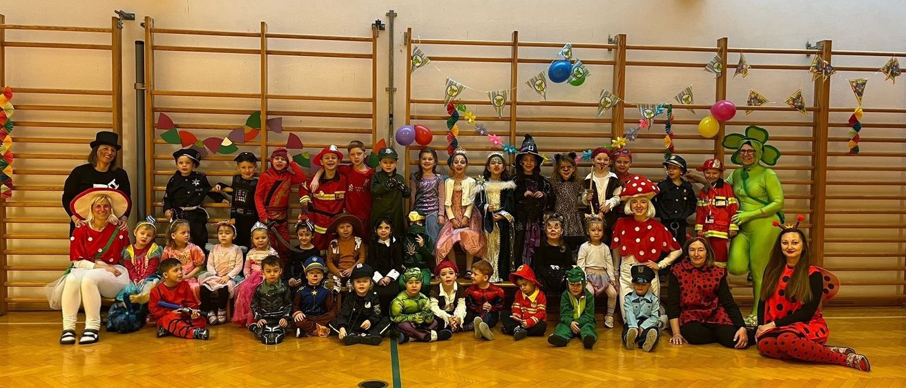 Eine Gruppe von Kindern in Kostümen posiert für ein Foto in einer Turnhalle. Hinter ihnen befinden sich Luftballons und Dekorationen.