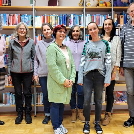 Eine Gruppe von Frauen und Männern steht vor einem Bücherregal und lächelt für ein Foto. Sie tragen alle Winterkleidung.