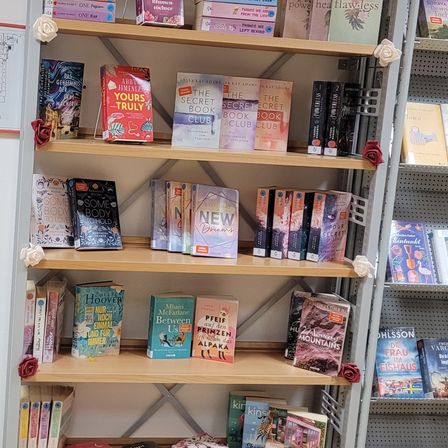 Ein Bücherregal mit verschiedenen Büchern, darunter Titel wie 'Der geheime Buchclub', 'Zwischen uns' und 'Einen Körper zum Halten'. Das Regal hat Holzplatten und ist mit roten Rosen dekoriert.