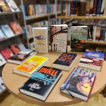 Ein Tisch zeigt mehrere Bücher, darunter Titel wie 'Die Liebe der Bücher,' 'Die junge Nacht,' 'Monsieur Itcômete' und 'Onkel.' Der Hintergrund zeigt ein Bücherregal mit vielen Büchern.