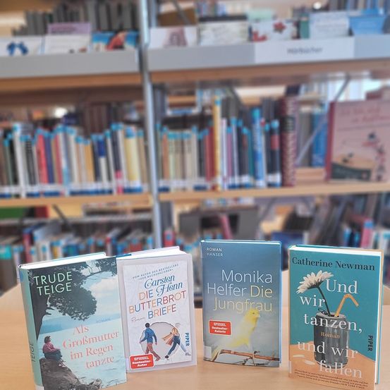 Vier Bücher sind auf einem Tisch in einer Bibliothek ausgestellt. Die Bücher tragen die Titel 'Als Großmutter im Regen tanzte', 'Die Butterbrot Briefe', 'Monika Helfer Die Jungfrau' und 'Und wir tanzen, und wir fallen'.