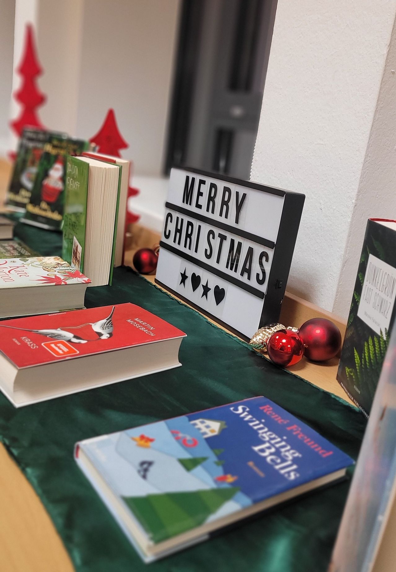 Eine Sammlung von Büchern auf einem Tisch, darunter ein rotes Buch mit dem Titel 'Krass', ein Weihnachtslicht-Schild mit der Aufschrift 'Frohe Weihnachten' und andere Bücher.