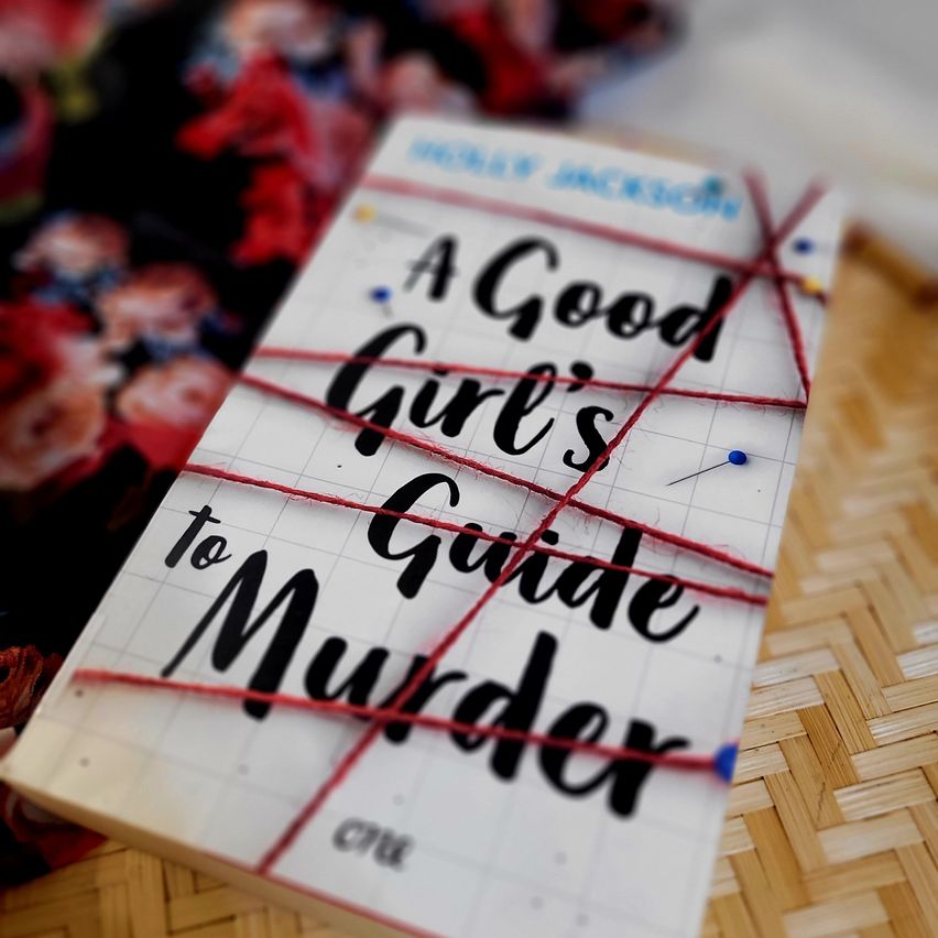 Ein Buch mit dem Titel 'A Good Girl's Guide to Murder' ist mit rotem Faden auf einem gewebten Untergrund befestigt. Der Buchdeckel hat ein Gittermuster.