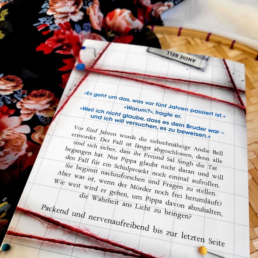 Eine Nahaufnahme einer Buchseite mit deutschem Text, mit rotem Faden angeheftet, auf einem Blumenhintergrund platziert. Der Text handelt von einem Mordfall und der Suche nach der Wahrheit.