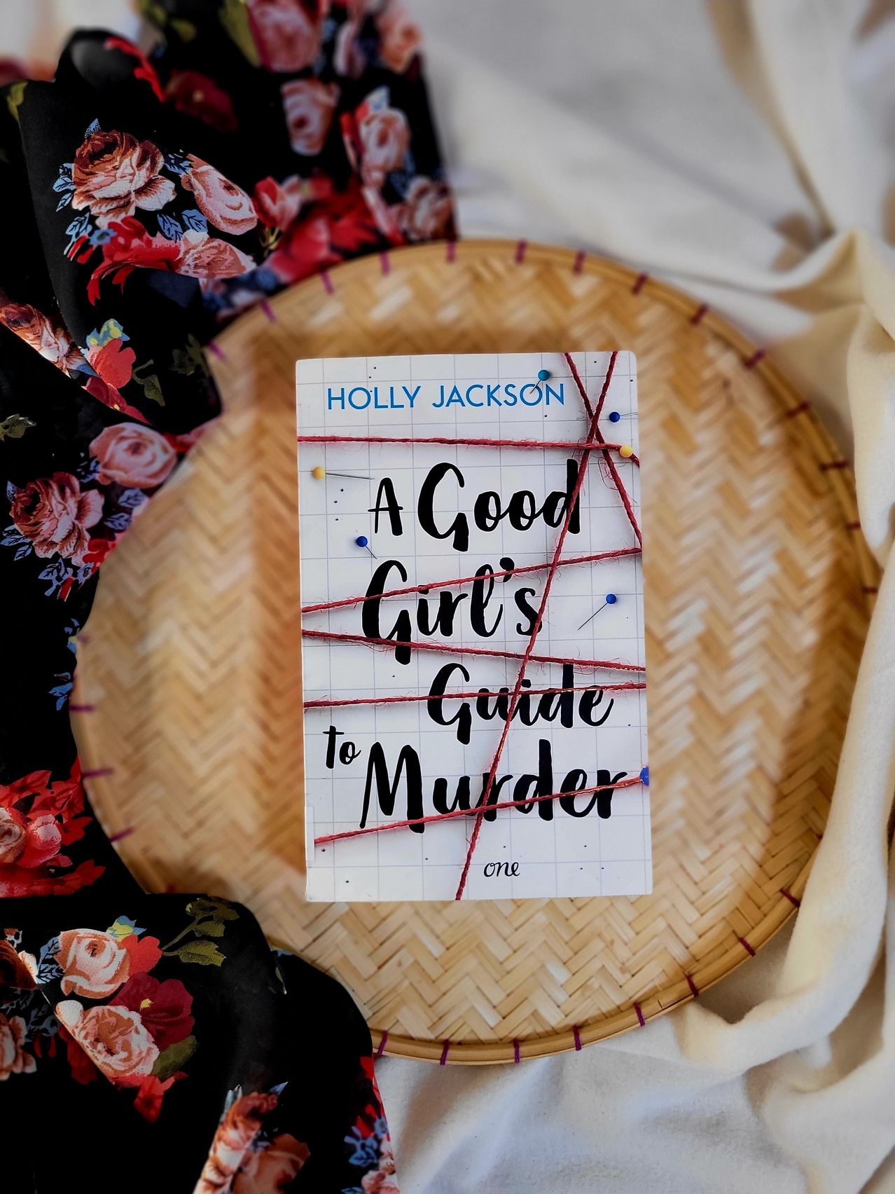 Ein Buch mit dem Titel 'Ein guter Leitfaden für Mädchen zum Morden' von Holly Jackson liegt auf einem geflochtenen Korb mit einem Blumentuch im Hintergrund.