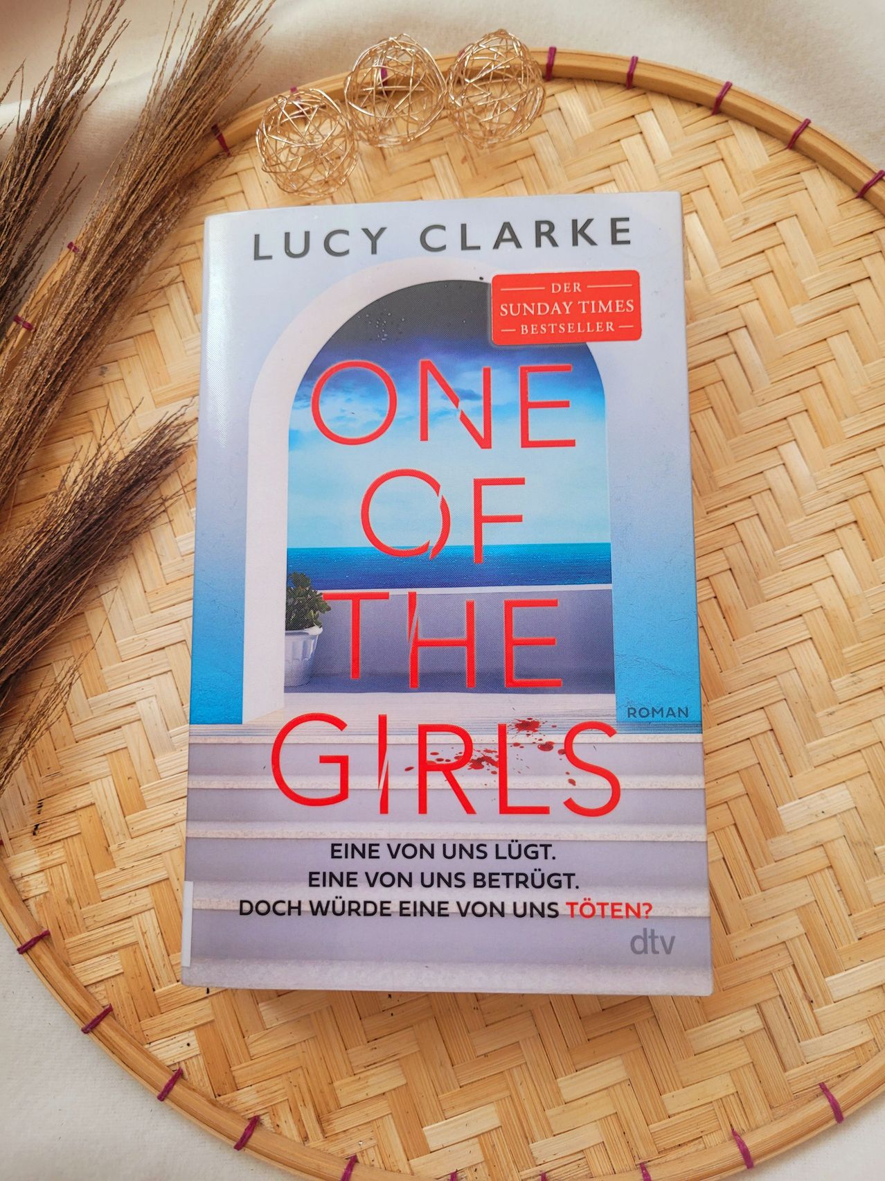 Das Buchcover von 'One of the Girls' von Lucy Clarke zeigt eine dramatische Szene mit Treppen zum Meer, betont durch rote Flecken, und einem Titel in kräftigem Rot.