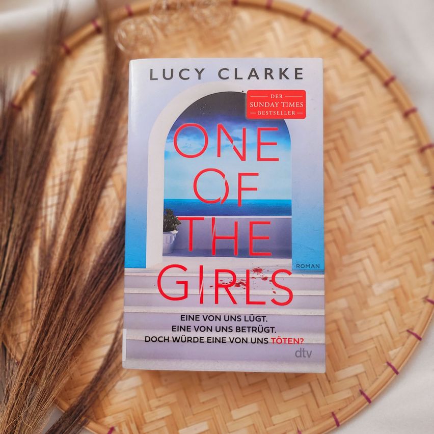 Das Buchcover von Lucy Clarkes 'One of the Girls' auf einem geflochtenen Tablett. Der Titel ist in fettem Rot, mit einer ruhigen Meereslandschaft und einem Haus im Hintergrund. Der deutsche Slogan lautet 'Eine von uns lügt. Eine von uns betrügt. Doch würde eine von uns töten?'.