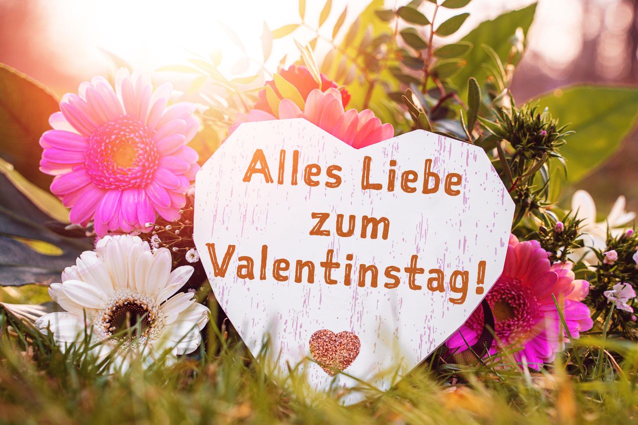 A heart-shaped sign with the message 'Alles Liebe zum Valentinstag!' is surrounded by flowers. The sign is white with orange text.