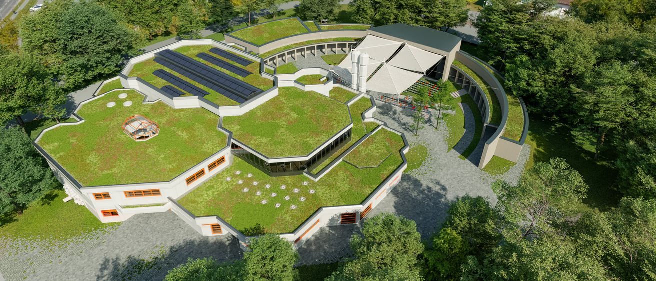Ein modernes sechseckiges Gebäude mit Solarmodulen auf dem Dach, umgeben von üppigem Grün und Bäumen, mit einem Parkplatz im Hintergrund.