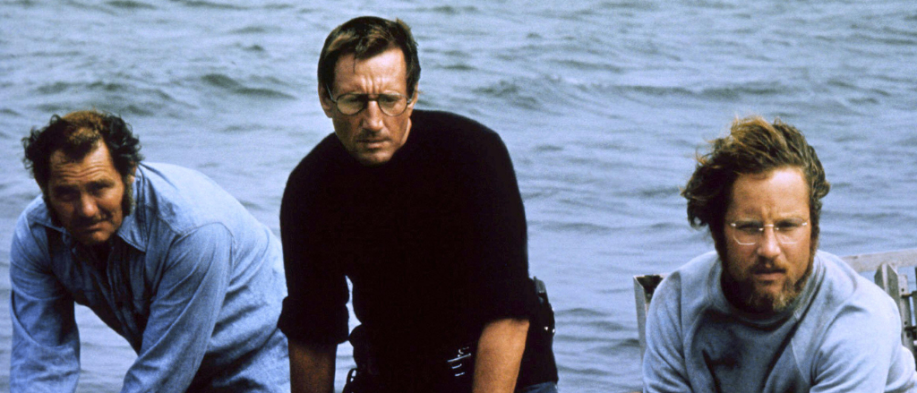 Drei Männer auf einem Boot. Ein Mann in einem schwarzen Shirt, einer in einem blauen Shirt und einer mit Bart, alle schauen in Richtung etwas vor ihnen. Das Boot ist auf dem Wasser.