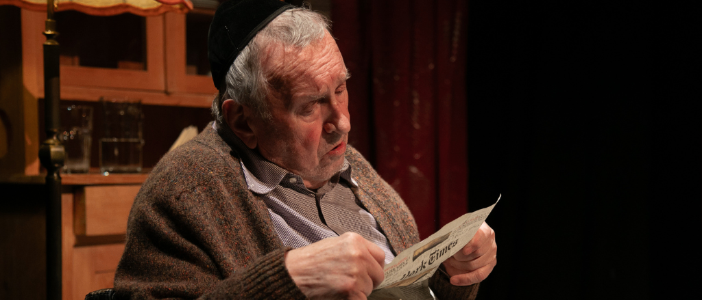 Ein älterer Mann mit schwarzer Kopfbedeckung sitzt auf einem Stuhl und liest eine Zeitung. Der Raum ist schwach beleuchtet, und hinter ihm steht ein Holzschrank.