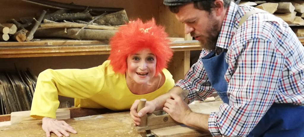 Ein Mann und eine Frau arbeiten in einer Werkstatt. Die Frau trägt ein gelbes Shirt und eine rote Perücke, während der Mann ein kariertes Hemd und einen Hut trägt. Sie lächeln beide und scheinen auf einer Holzwerkbank zu arbeiten. Hinter ihnen stehen Holzscheite auf den Regalen.