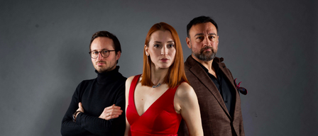 Drei Personen stehen vor einem grauen Hintergrund. Eine Frau in einem roten Kleid steht zwischen einem Mann mit Brille und einem Mann in einem braunen Anzug.