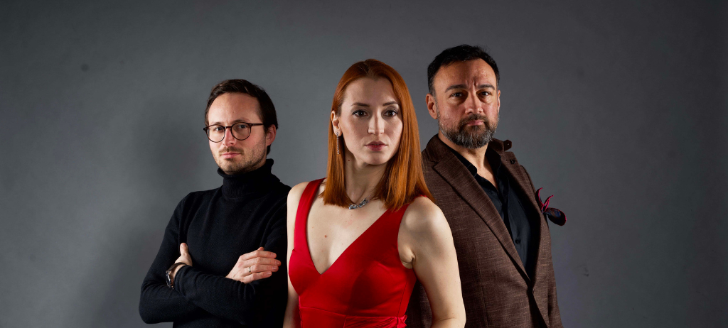 Drei Personen stehen vor einem grauen Hintergrund. Eine Frau in einem roten Kleid steht zwischen einem Mann mit Brille und einem Mann in einem braunen Anzug.