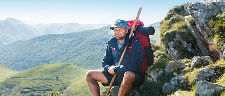 Ein Mann in blauer Jacke und Hut sitzt auf einem felsigen Berg mit Blick auf grüne Hügel und blauen Himmel. Er hält einen Stock und hat einen Rucksack auf.